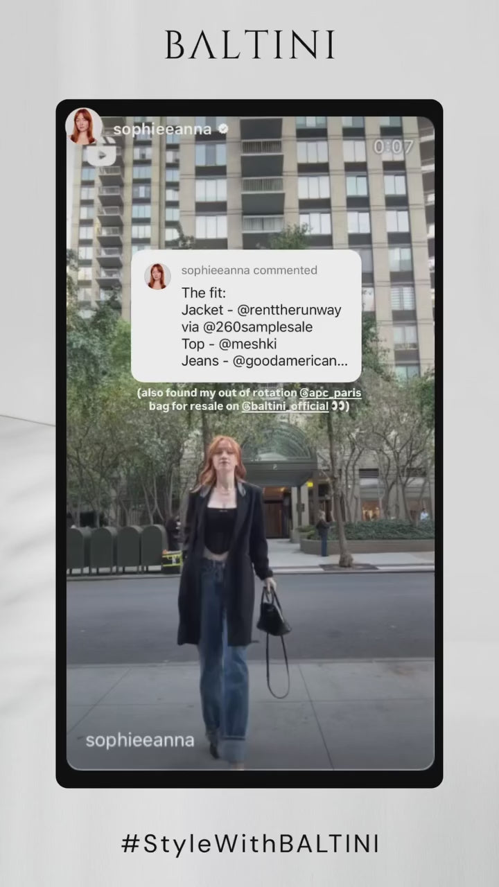 A fashionable woman walks confidently in an urban environment, carrying a bag. She wears a long black coat, a black top, and blue wide-leg jeans. The background features a tall modern apartment building with greenery. the bag for resale on @baltini_official. The BALTINI branding and the hashtag ‘#StyleWithBALTINI’ are displayed at the bottom of the video.