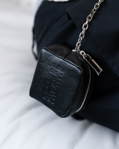 Close-up of a small black leather pouch attached to a silver chain and a black fabric strap. The pouch has embossed text reading “MM DESIGNER” and a small circular logo beneath the text. The background is soft white fabric.