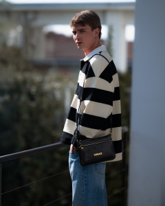 A young man stands outdoors, leaning on a railing. He wears a black and white striped long-sleeve shirt and blue jeans. He carries a small black crossbody bag.