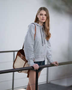 A young woman with long blonde hair stands outdoors, resting her hand on a railing. She wears a grey hoodie and carries a medium-sized handbag with brown leather accents and beige woven texture.